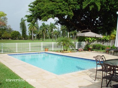 Pale square pavers surround a small square pool beside metal dining furniture, a white garden fence and trees.