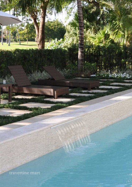 A long pale coping ledge tops a raised pool wall with a narrow sheet of falling water and loungers on lawn beyond.