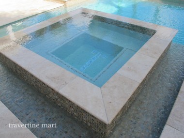 An angled view shows light stone coping meeting at the corners of a square mosaic-faced spa.