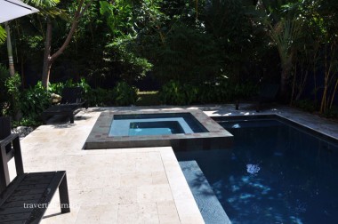 Pale square paving borders a pool and raised square spa beside dense tropical planting and dark outdoor furniture.