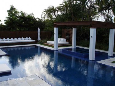Pale square paving surrounds a dark-blue pool beside a white-column pergola, loungers and dense palms.
