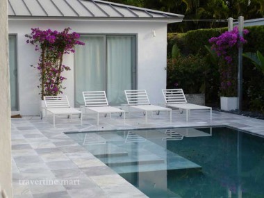 Pale square paving borders a reflective pool beside white chaise lounges, a sliding door and purple flowering vines.