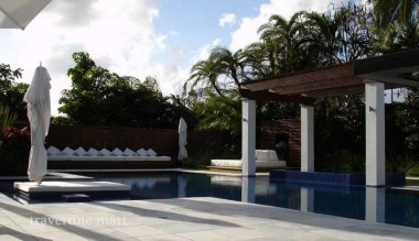 Pale square paving runs beside a blue pool beneath a timber pergola with white columns and cushioned seating.