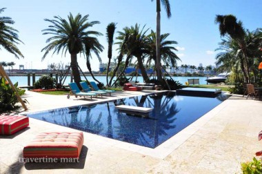 Light square pavers surround a long blue pool beside palms, open water and low lounge seating with red cushions.