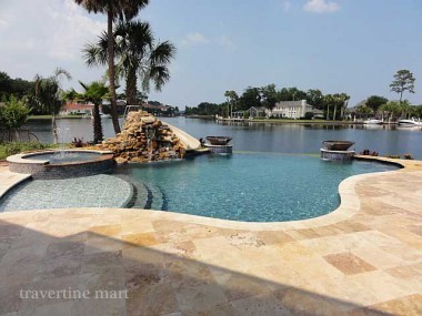 Warm cream and gold square pavers surround a freeform waterside pool with a raised spa and rock water feature.