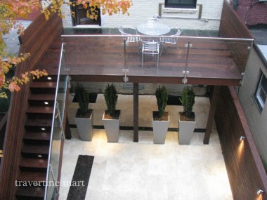 Pale rectangular stone paving forms a lower courtyard beneath an elevated wood dining terrace with glass railings and tall planters.
