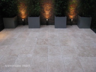 Pale rectangular pavers extend toward tall planters against an illuminated brick wall in an evening courtyard view.