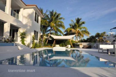 Pale square pavers surround a waterside pool beside a house, palms, low seating and stretched fabric shade sails.