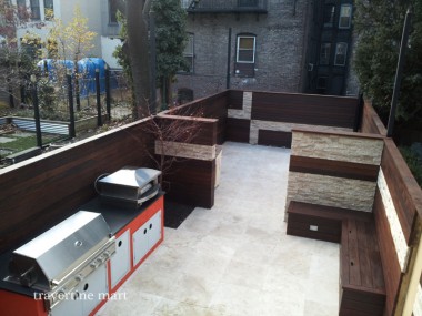 Pale square pavers cover an enclosed courtyard with cooking appliances, built-in timber benches and a small tree.