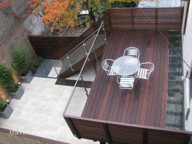 An overhead view shows pale stone paving below an elevated wood deck with a round dining table, glass railings and stairs.