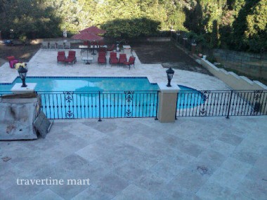 Large stone pavers cover a broad upper terrace overlooking a fenced rectangular pool and a courtyard furnished with red chairs.