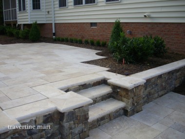 A short run of pale stone steps passes through a capped stacked-stone retaining wall beside a house patio.