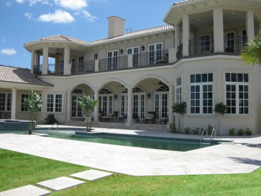 Light rectangular pavers form a broad deck around a long pool in front of a house with arches and an upper balcony.
