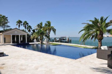 Light rectangular pavers surround a blue waterside pool with palms, large planters and a small pool house beyond.