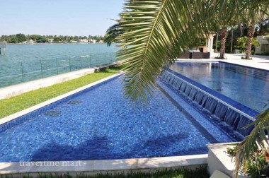 Light rectangular coping edges a blue pool with a long tiled spillover wall beside palms and waterside lawn.