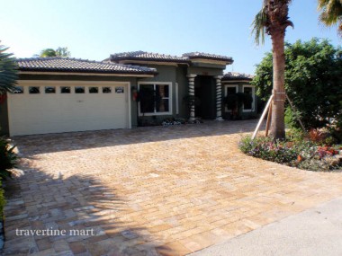 Small golden stone pavers cover a driveway in front of a tiled-roof house and double garage, bordered by palms and planting.