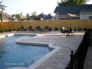 Brown rectangular pavers surround a pool with a curved projecting deck, outdoor dining furniture and a timber fence.