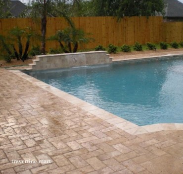 A close view shows brown rectangular pavers and pale coping meeting at a pool corner below a planted water-feature wall.