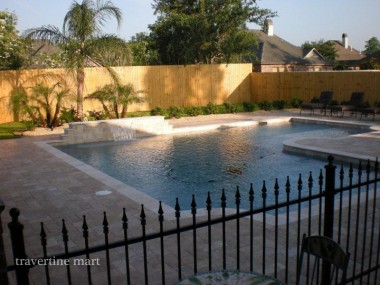 Brown pavers surround a rectangular pool with a low rear water-feature wall, palms and a timber privacy fence.