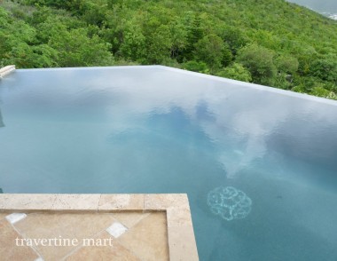 Rectangular stone pavers form an angular edge beside a pool where the water extends toward dense green planting.