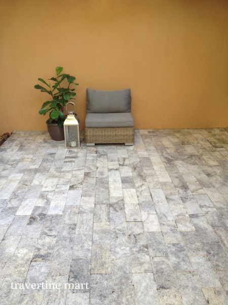 Gray and cream rectangular pavers cover a small seating area with a cushioned chair and a potted plant against an ochre wall.