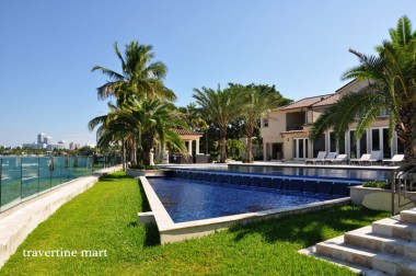 Light coping edges a blue pool above a lawn, with palms, glass fencing and open water beyond.