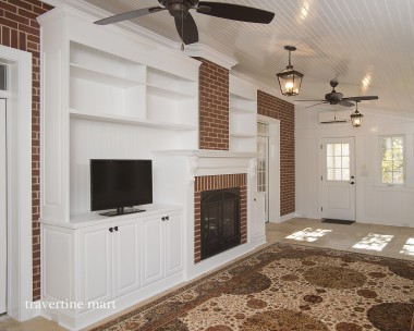 A cream mixed-size tile floor runs beside a patterned rug in a white porch with a brick fireplace and ceiling fans.