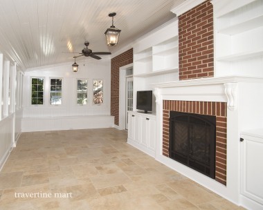 Cream mixed-size tiles cover a bright enclosed porch with white cabinetry, ceiling fans and a brick fireplace.