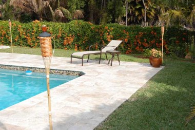 Pale mixed-size paving surrounds a pool beside a chaise lounge, flowering hedges, palms and a garden torch.