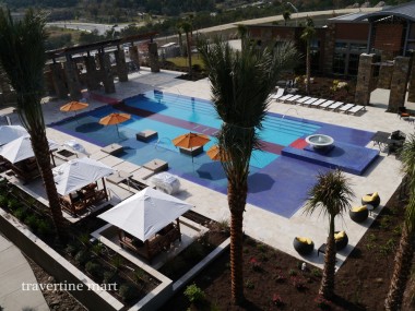 An overhead view shows a pool courtyard with pale paving, blue pool water, palm trees, umbrellas and shaded lounge seating.