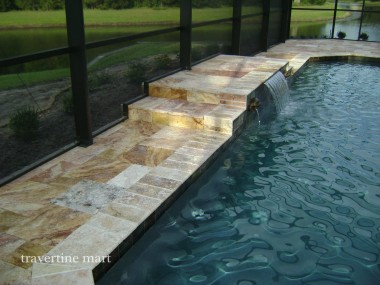 Warm mixed-size paving borders a pool within a screened enclosure, with a raised stone section spilling water into the pool.