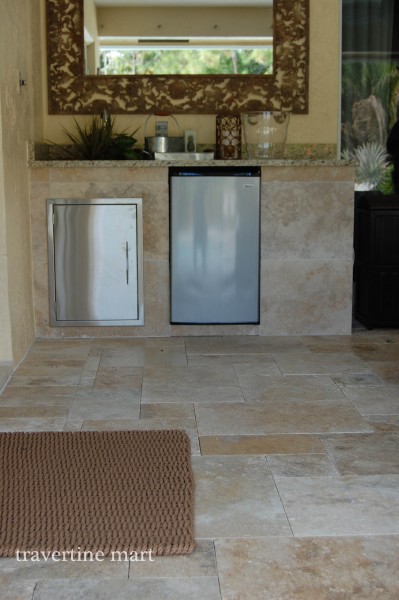 Mixed-size pale stone paving extends beneath a covered bar with metal cabinets, a sink and a large framed mirror.