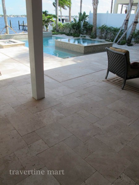 Pale mixed-size pavers cover a columned terrace overlooking a pool, raised square spa and open water.