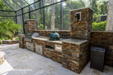 Pale mixed-size pavers cover the floor beneath a stone-faced outdoor kitchen with metal cabinets and cooking appliances.