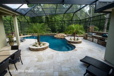An angled view across pale paving shows a screened pool, palm planters, woven lounge seating and a stone-faced outdoor kitchen.