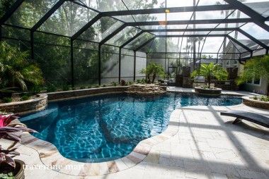 Pale mixed-size paving follows a freeform pool inside a screened enclosure with palms and planted raised beds.