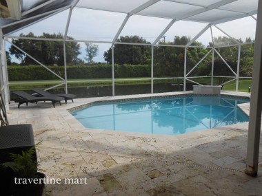 Cream and brown mixed-size pavers surround a blue pool inside a screened enclosure overlooking a green lawn.