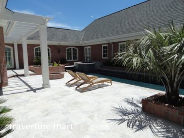 Pale gray and cream paving surrounds a pool beside a brick house, a white pergola and wooden chaise lounges.
