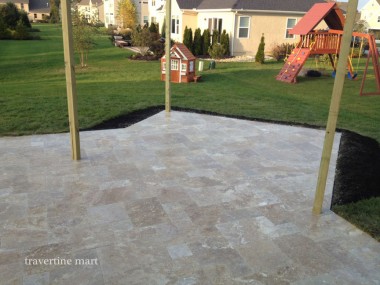 Pale mixed-size paving extends between timber pergola posts toward a lawn with a play structure beyond.