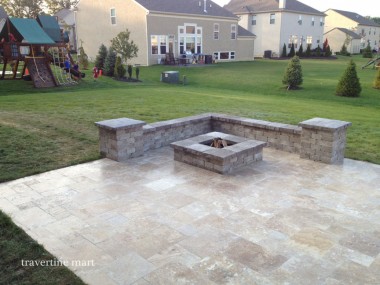 A pale mixed-size paved court surrounds a square fire pit and low stone seating walls beside an open lawn.