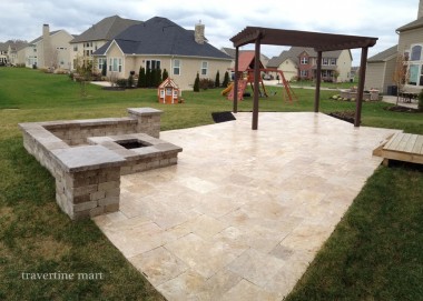Pale mixed-size paving forms a backyard terrace with a timber pergola, a built-in stone fire pit and lawn beyond.