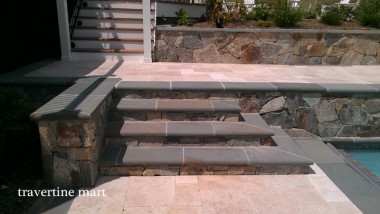 Broad dark-edged steps join two pale stone terraces beside a pool and a stacked-stone wall.