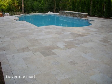 A broad expanse of pale mixed-size paving surrounds a blue pool with a rounded end and a low stone wall.