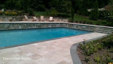 A light stone pool deck follows a curved planting bed below a retaining wall topped with outdoor chairs.