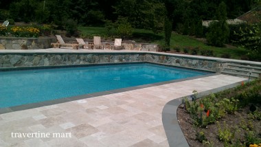 Pale pavers curve around a planted bed beside a blue rectangular pool and a stone retaining wall.