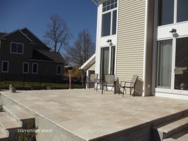 A raised stone patio runs along tall house windows, with a broad edge step and metal chairs near the doors.