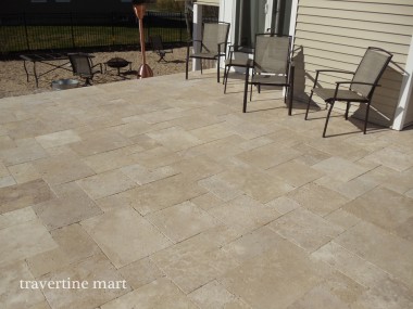 A broad mixed-size stone patio holds metal chairs and a small table beside light-colored house siding.