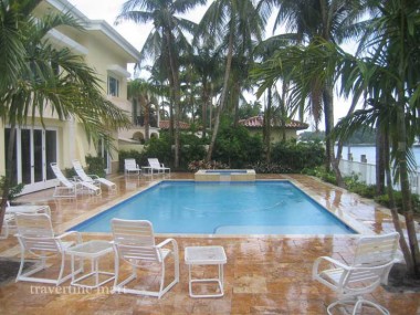 Golden paving surrounds a rectangular blue pool with white lounge chairs, palms and a covered house terrace.