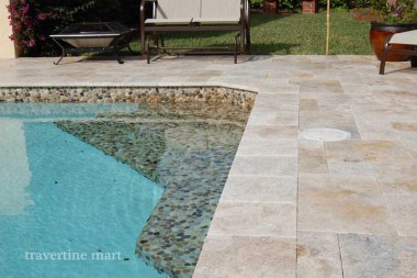 Pale mixed-size pavers meet stone coping beside a curved mosaic-lined shallow pool ledge.