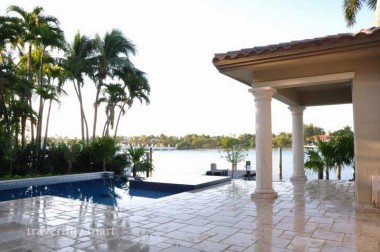 Brown mixed-size tiles cover a columned terrace beside a pool, with palms and open water beyond the lawn.